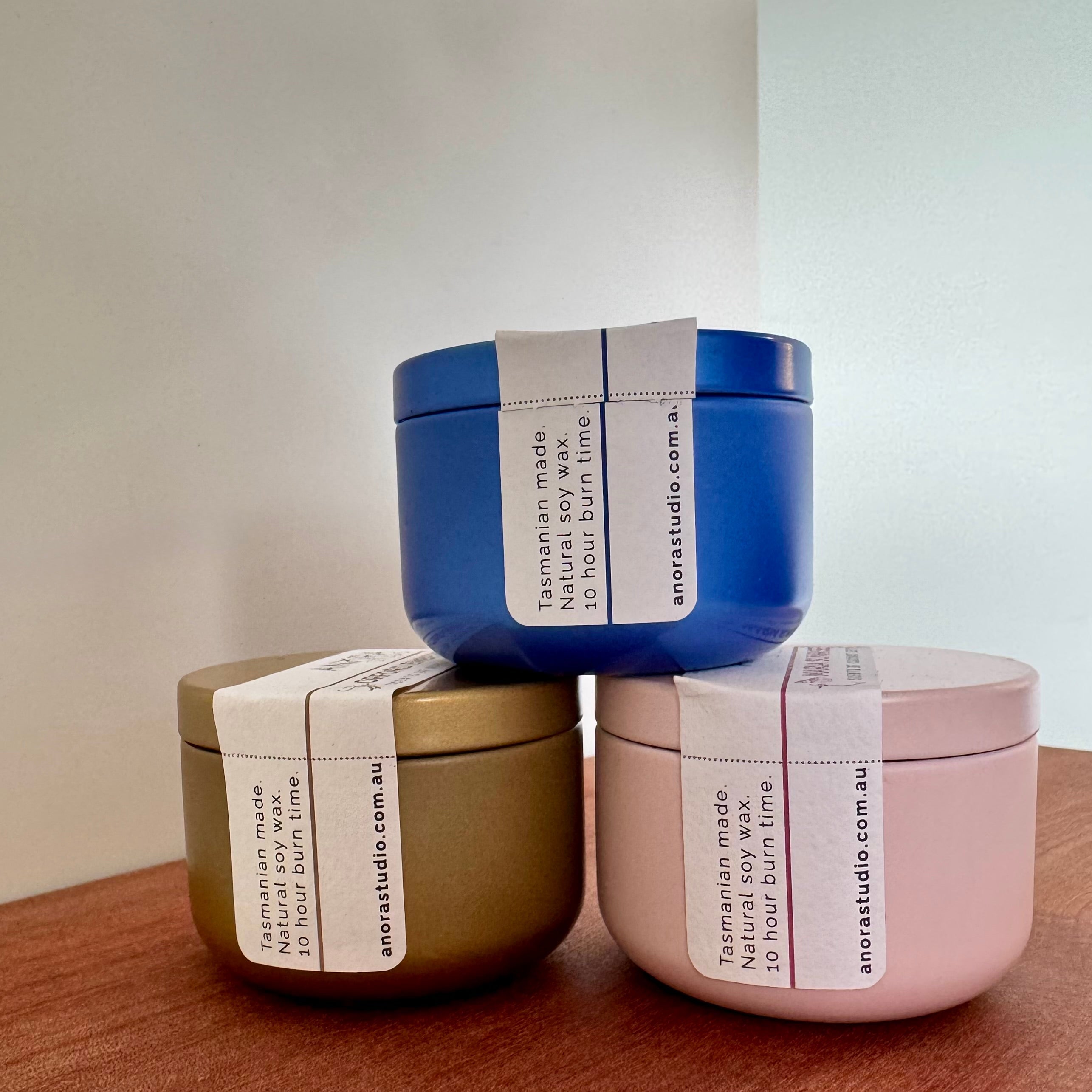 Three round candle tins in 3 different colours stacked in a pyramid on a wooden board, with a white label reading 'anorastudio.com.