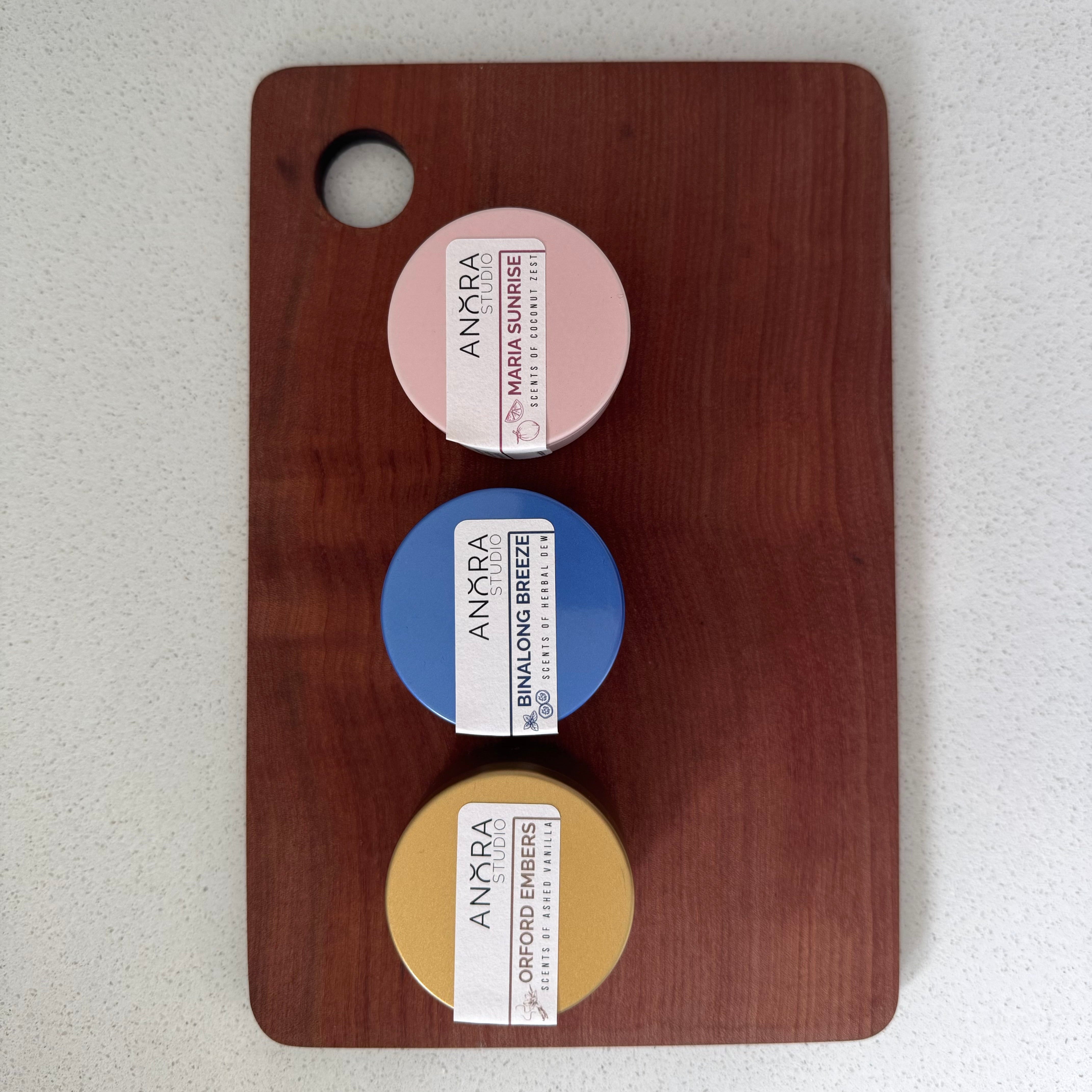 Three round candle tins from the brand ANORA, arranged vertically on a dark brown wooden cutting board. The jars are labeled 'MARIA SUNRISE' (pink), 'BINALONG BREEZE' (blue), and 'ORFORD EMBERS' (yellow), set against a light gray speckled countertop.