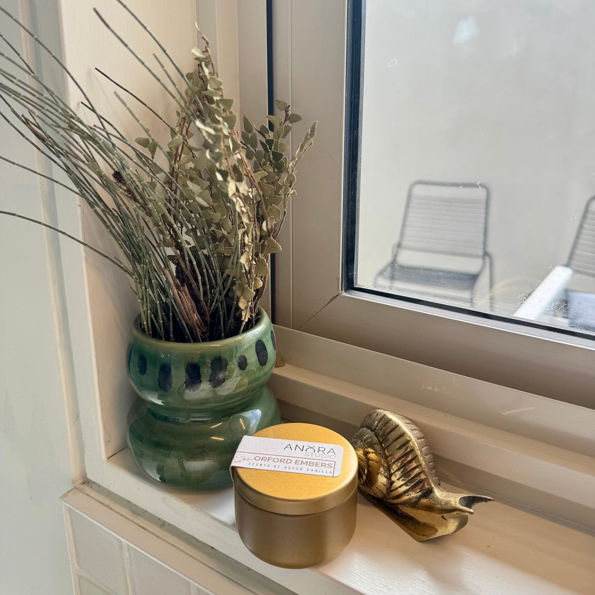 A round gold tin candle with a white and black label sits on a windowsill next to a green ceramic vase with dried leaves and a small gold decorative gold snail. The window shows an outdoor patio chair outside.

