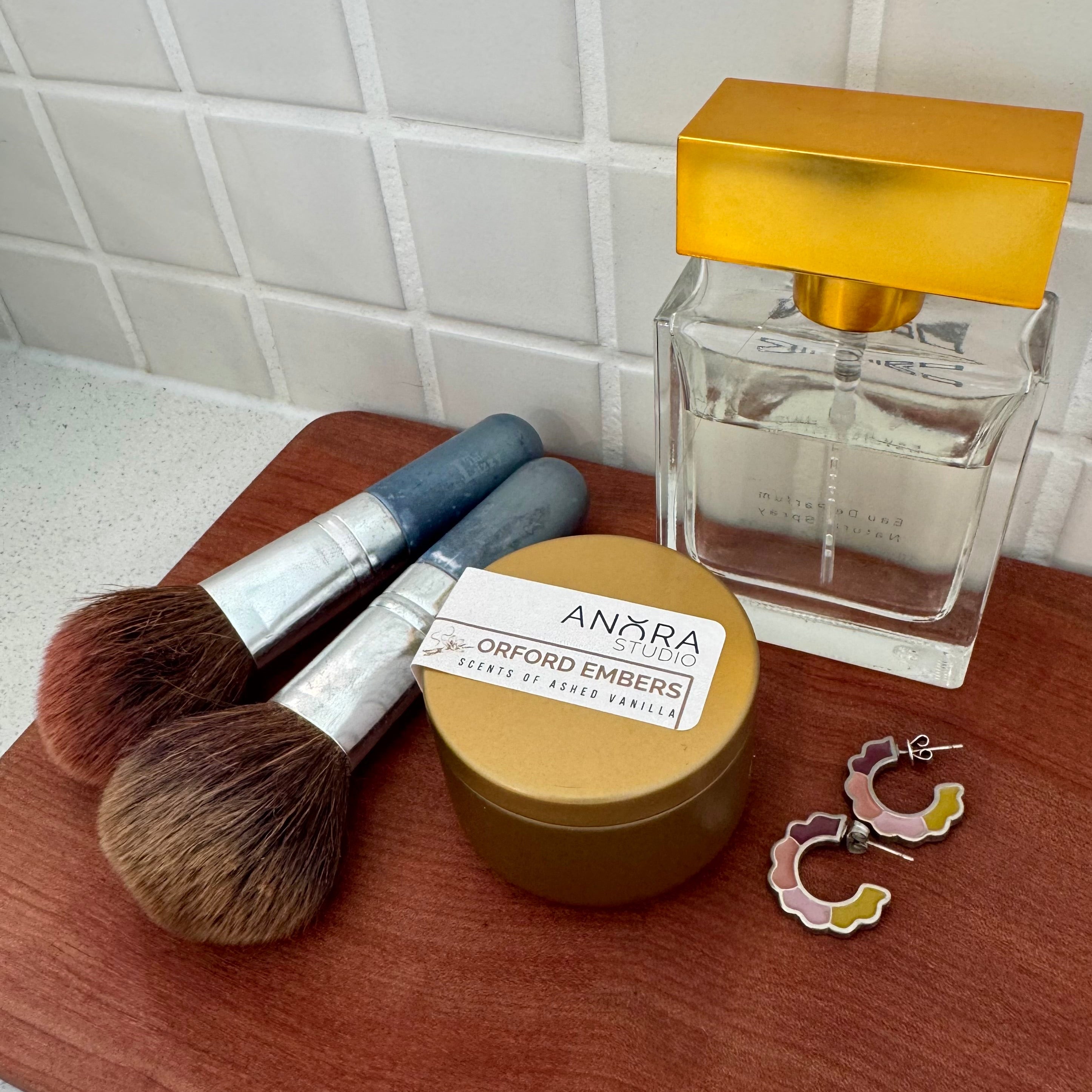 A styled flat-lay of Anora Orford Embers candle in a small metal  round gift tin on a wooden board, also featuring a perfume bottle, two makeup brushes, and two small earrings against a white tiled background.