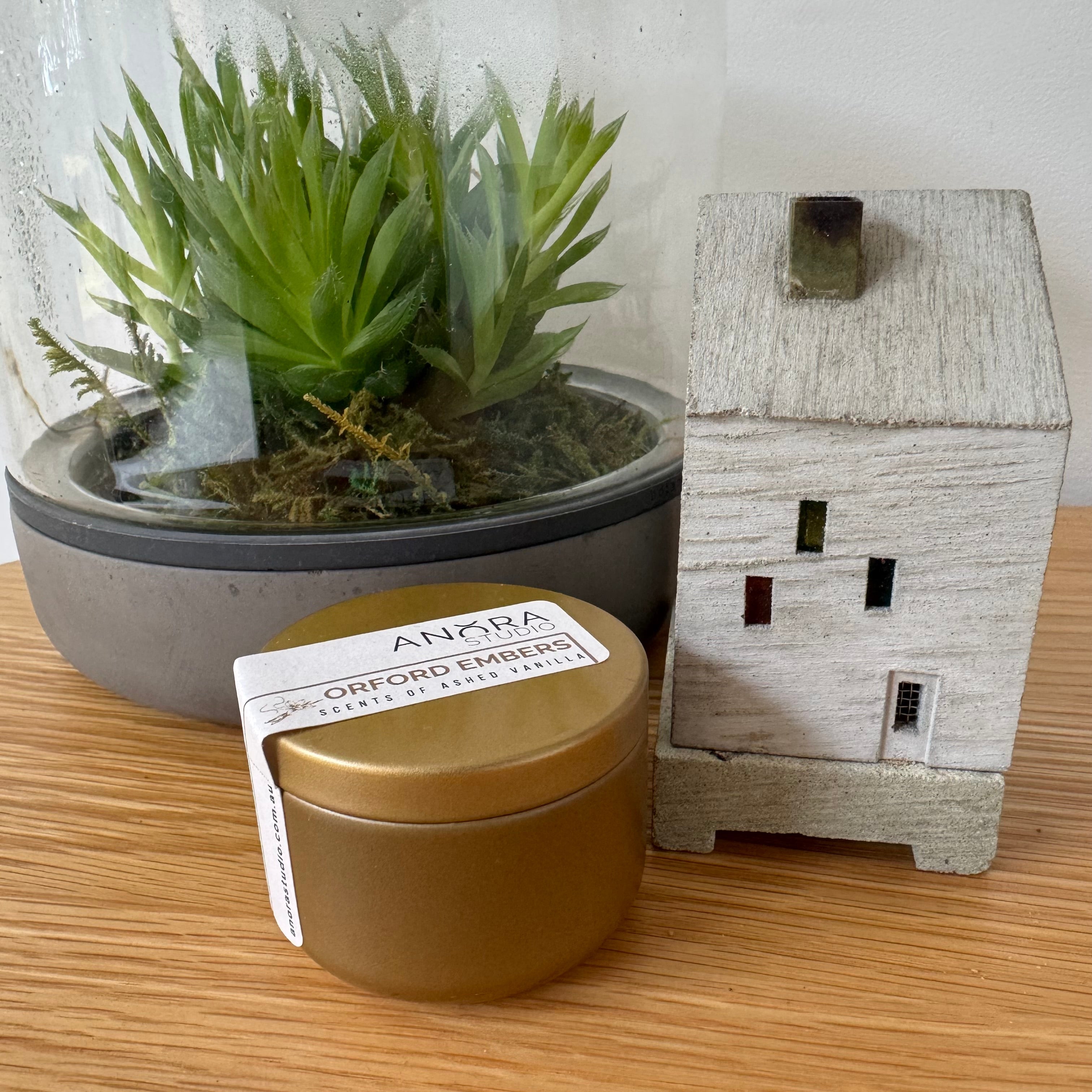 A gold tin candle with a paper label reading 'ANORA ORFORD EMBERS' sits on a wooden surface next to a small white decorative house and a glass dome terrarium with a green succulent.