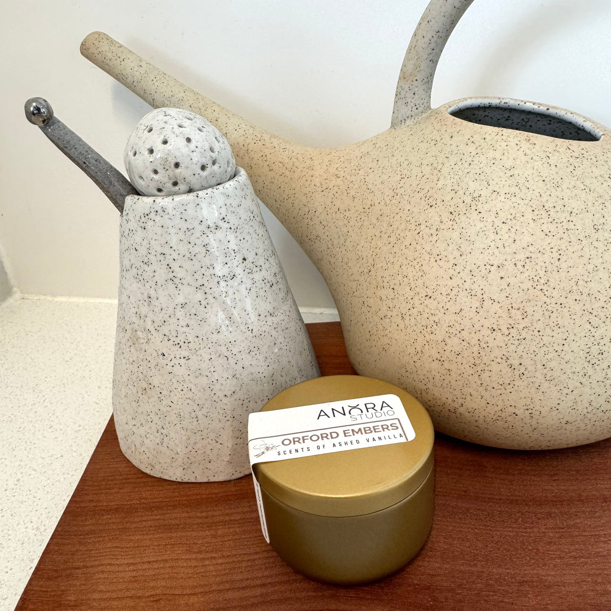A small round gold tin of Anora Orford Embers candle tin sits on a wooden cutting board, styled with ceramic kitchenware in the background.